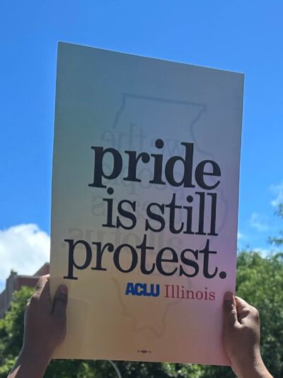 hands holding up sign with rainbow background and gray text "pride is still protest"