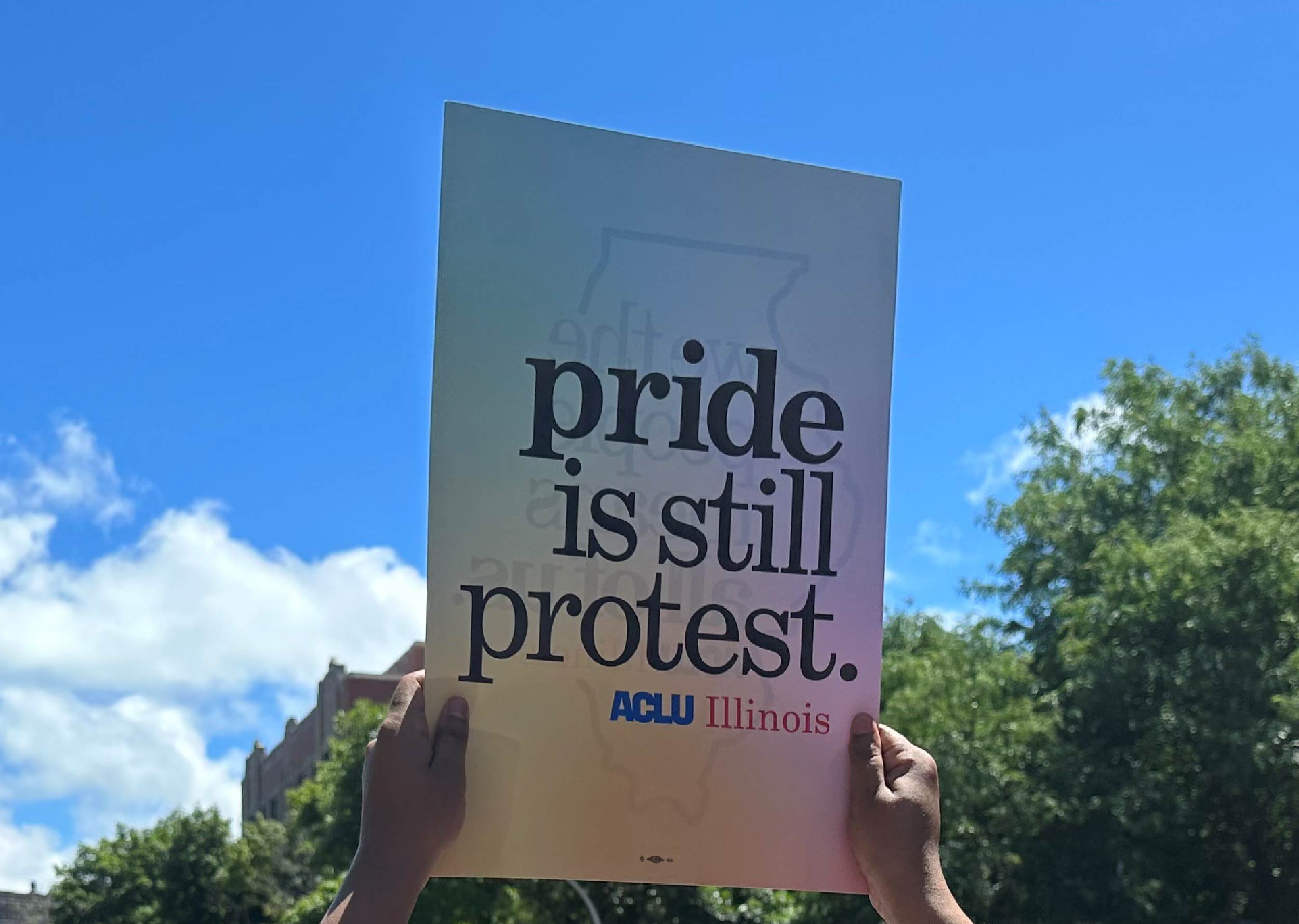 hands holding up sign with rainbow background and gray text "pride is still protest"