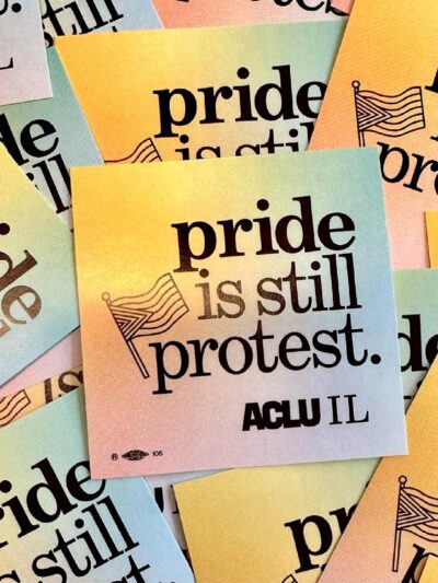 Photo of pile of stickers spread out. Stickers have a rainbow background and say "pride is still protest" with an outline of a Pride Flag