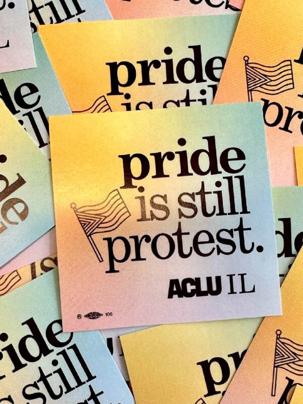 Photo of pile of stickers spread out. Stickers have a rainbow background and say "pride is still protest" with an outline of a Pride Flag