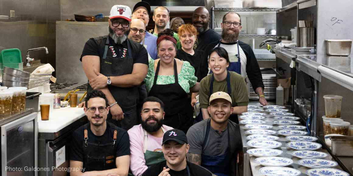 Group photo of chefs and staff who helped during our chef event pose in the kitchen with a row of plates to their right