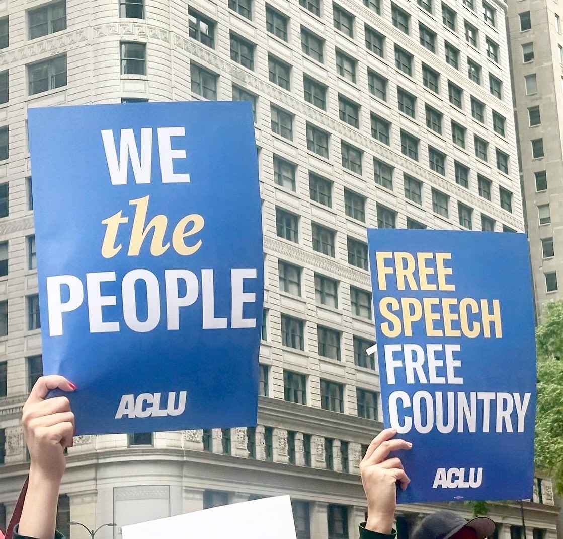Hands holding up blue signs with white text. One sign says "We the people" and one says "free speech, free country"