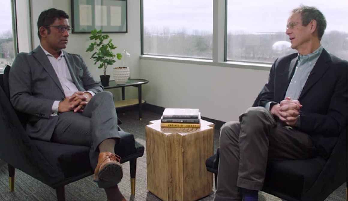 Keynote speakers Aziz Huq on the left and David Cole on the right sit in chairs facing each other with a table of books between them and a background of windows looking out over a park